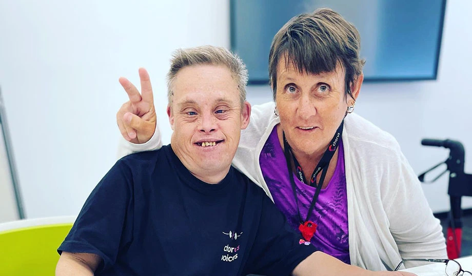 Two people sitting together at a table, smiling at the camera, with one person playfully making a peace sign during a community event